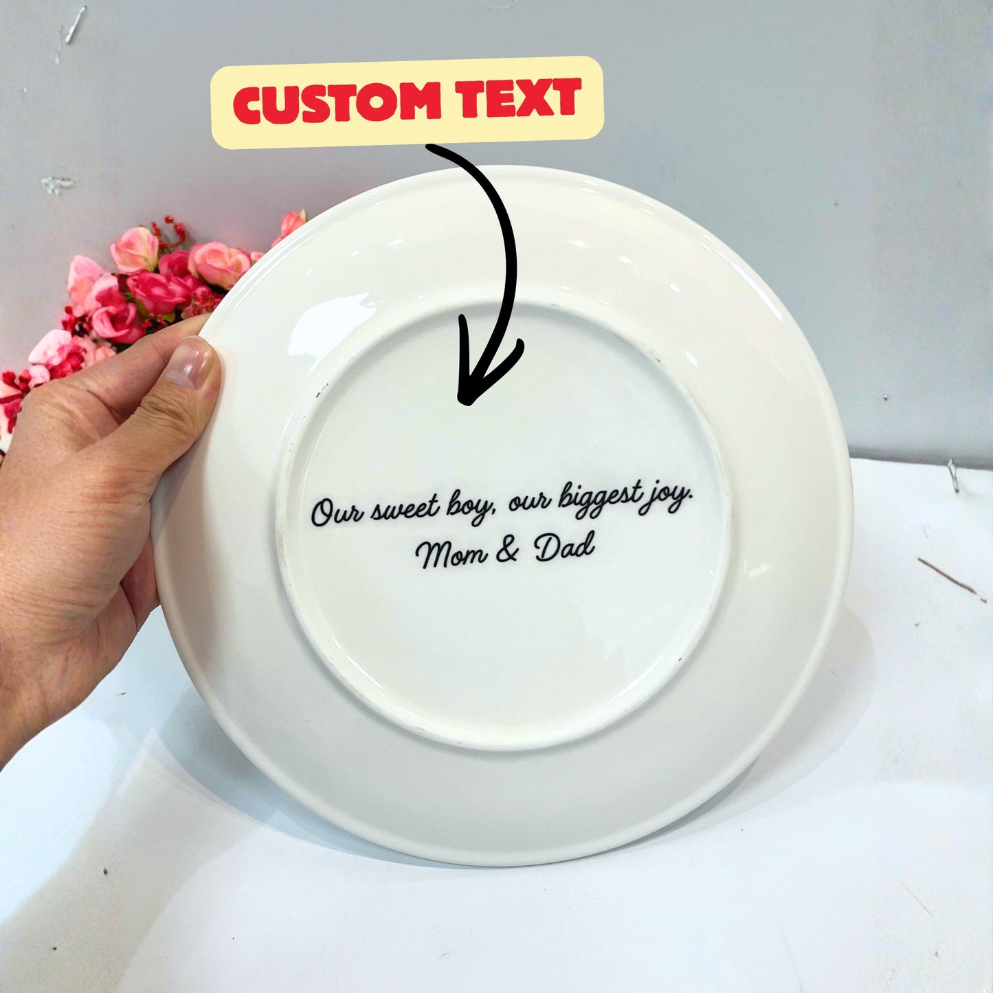 White plate with personalized text held by a hand, with flowers in the background.